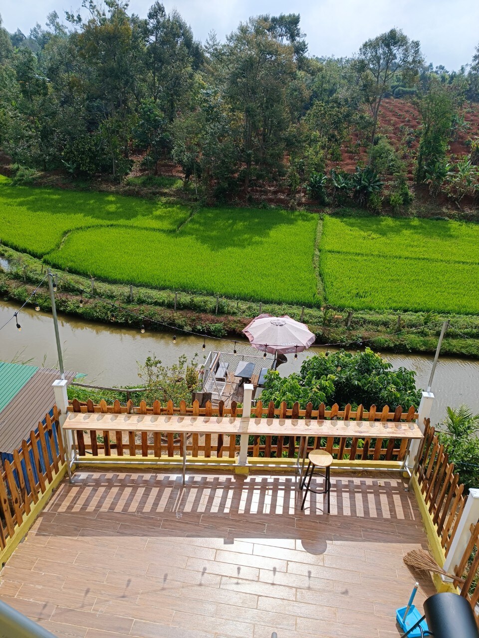 A balcony view overlooks a tranquil canal bordered by lush green rice fields and dense trees. A patio umbrella is visible near the water's edge, while a wooden barstool is positioned on the balcony, inviting relaxation amidst the serene landscape.