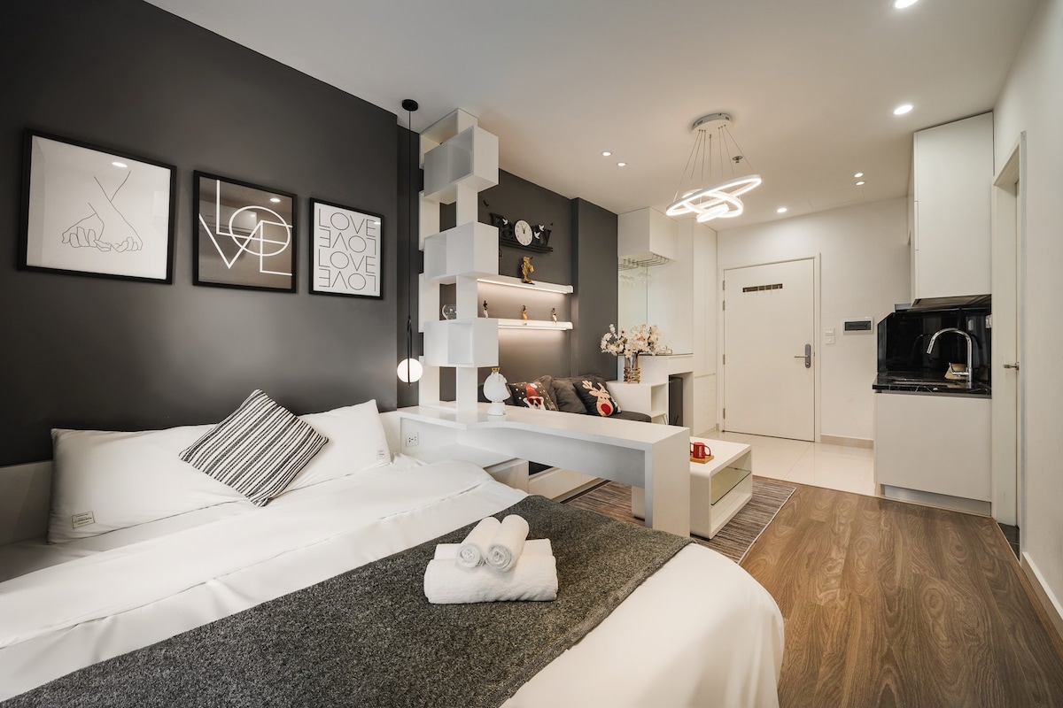 A modern room is featured with a spacious bed covered in crisp white bedding and a gray throw. Decorative art pieces adorn the dark walls. A stylish partition with shelves separates the sleeping area from a small sitting area with cushions, creating a cozy ambiance.