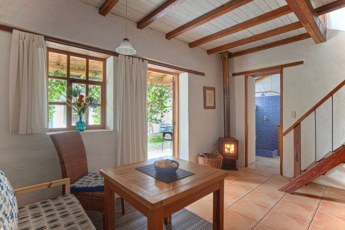 A cozy living area is highlighted by wooden beams and tiled flooring. A wooden table is centered between a chair and a sofa, with a small vase of flowers atop it. A wood stove provides warmth, while light fills the space through two windows, showcasing views of the outdoor garden.