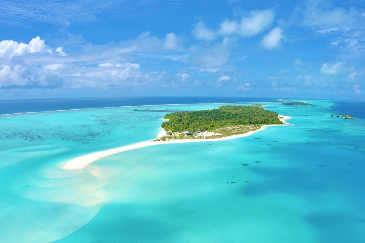 An aerial view of a private island showcases pristine white sandy beaches and a clear turquoise lagoon. Lush greenery is visible, and the surrounding waters feature gentle waves, creating a serene coastal environment.