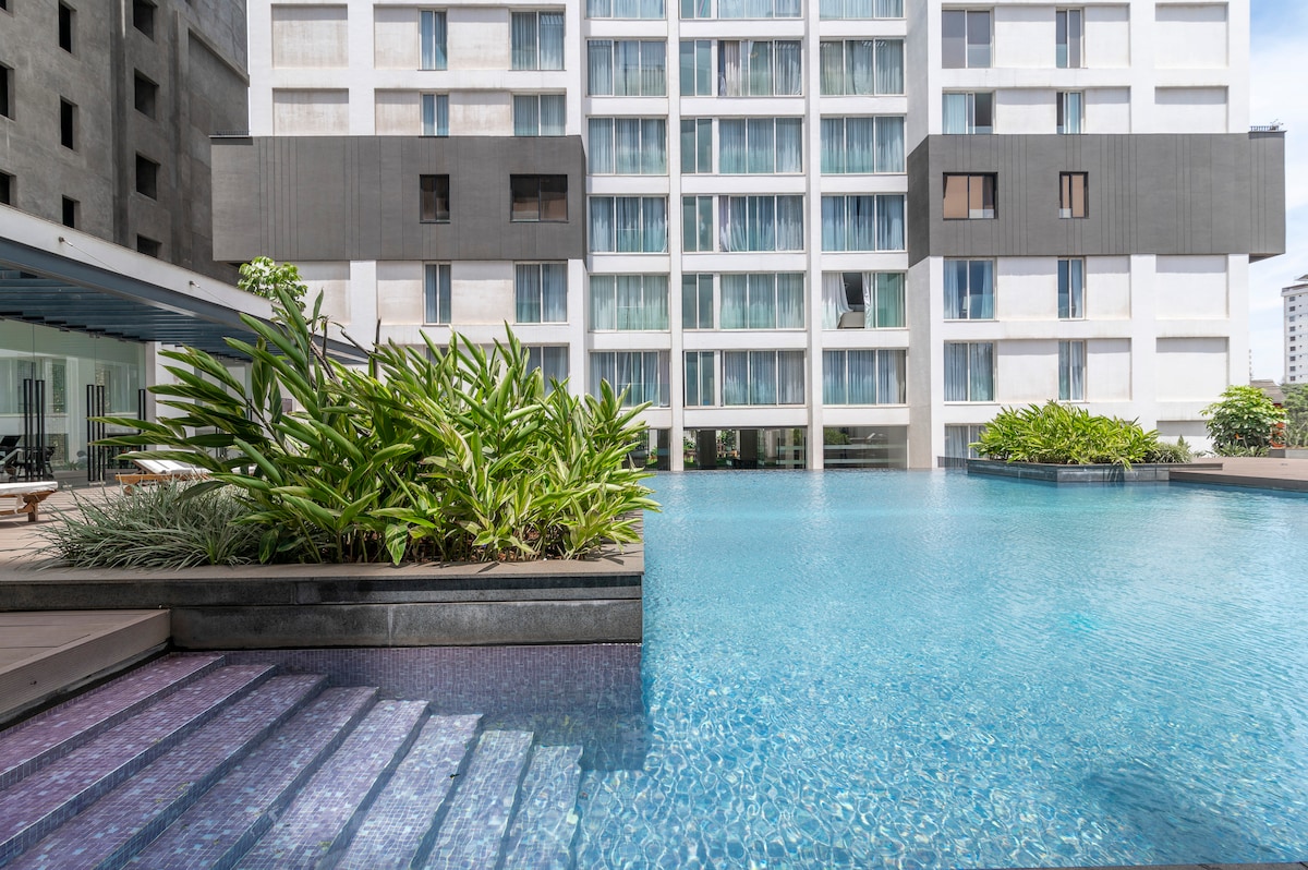 A large outdoor swimming pool reflects sunlight, surrounded by lush greenery. Modern architecture lines the background, while gentle steps lead from the pool's edge. The serene environment offers a blend of relaxation and modern design.