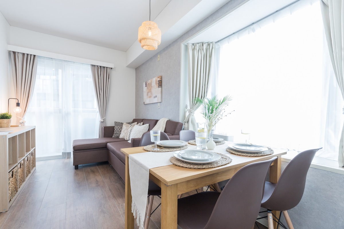A bright living and dining area features a spacious couch and a dining table set for four. Natural light pours in through large windows, enhanced by soft curtains. Neutral tones and simple decor create a calm ambiance, making the space inviting for relaxation and meals.