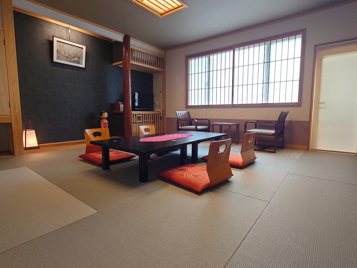 A spacious room features tatami flooring with a low table surrounded by several floor cushions. Natural light is filtered through large windows with traditional wooden frames, creating a serene atmosphere. Two armchairs are positioned beside the table, enhancing the inviting space.