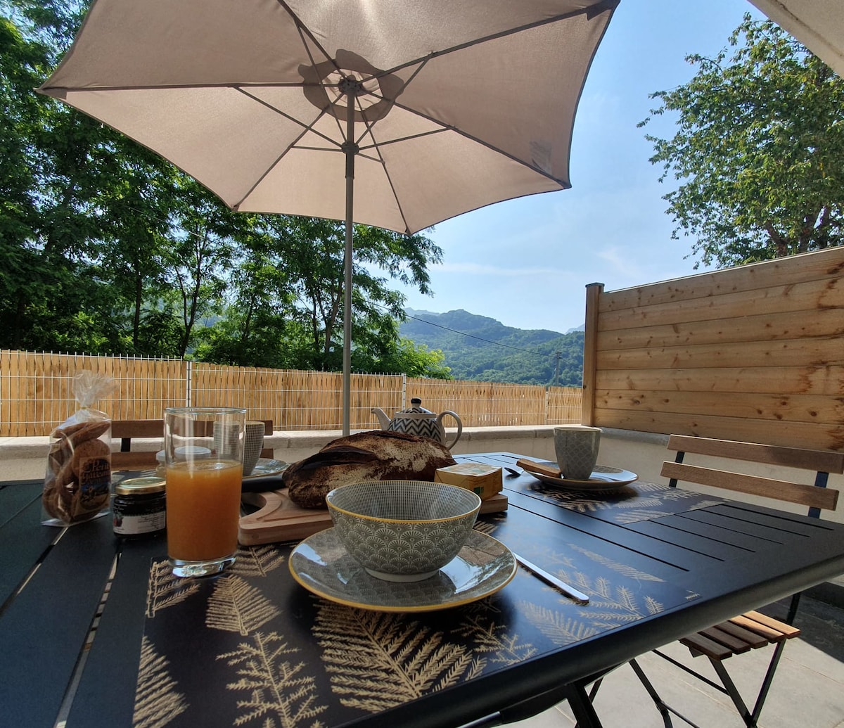 A terrace dining table is set with delicate plates and glasses, showcasing breakfast items. An umbrella provides shade, while scenic mountain views are visible in the background. Lush green trees add to the serene outdoor atmosphere.