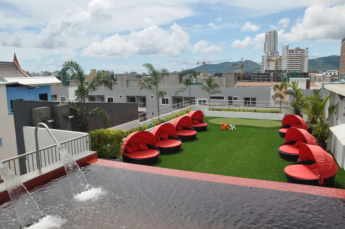 A rooftop area features a serene swimming pool with a cascading water feature, surrounded by lush green grass. Several circular red loungers are arranged thoughtfully around the space, offering comfortable seating with views of the city skyline and surrounding buildings.