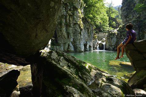 A serene water pool is surrounded by towering rocky cliffs and lush greenery. Sunlight reflects off the water's surface, creating a calm ambiance. The image also features a person sitting on a large rock, contributing to the tranquil setting.
