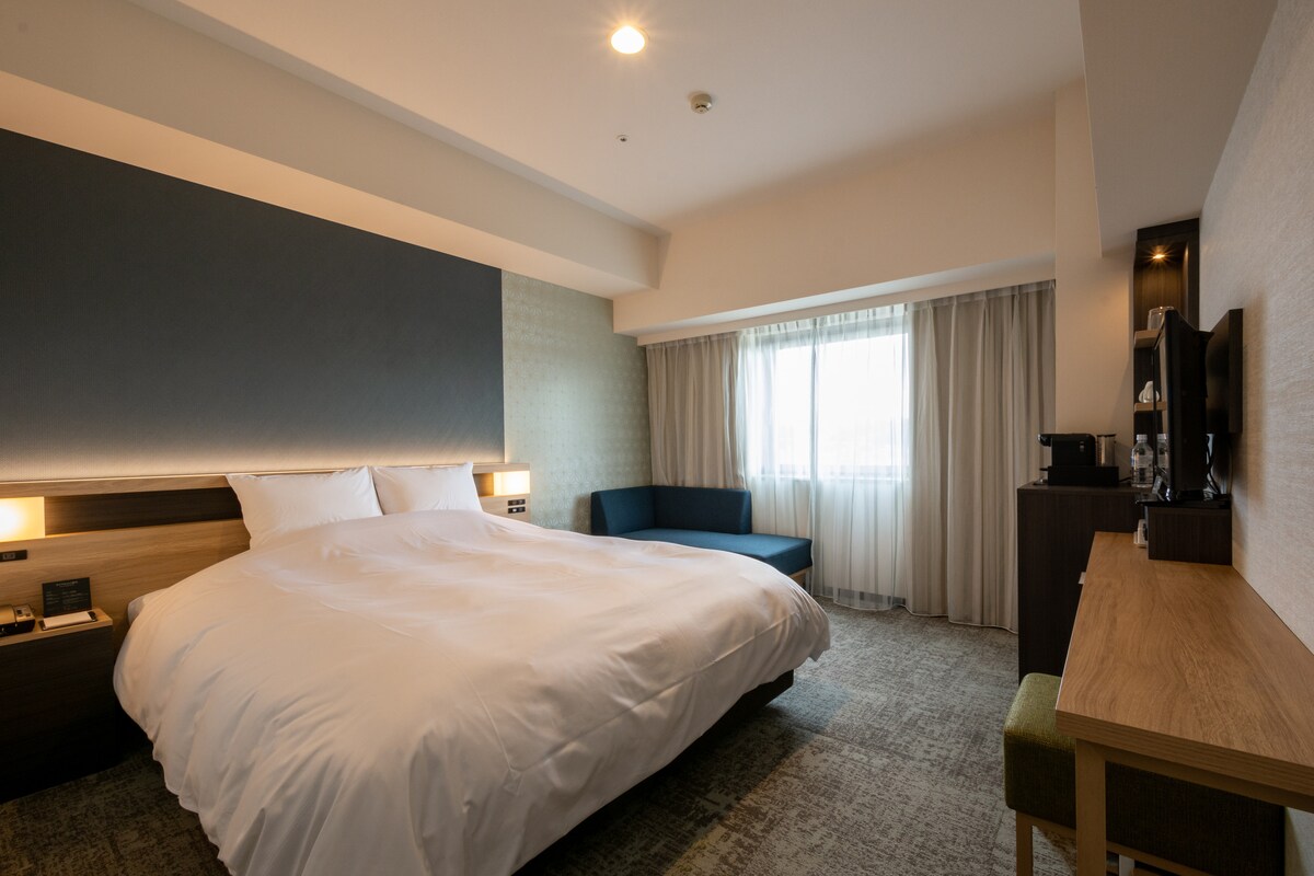 A spacious bedroom features a large bed with soft linens and a dark accent wall. Natural light enters through a window, complemented by sheer curtains. A comfortable seating area and a work desk with a television are also included in the room.