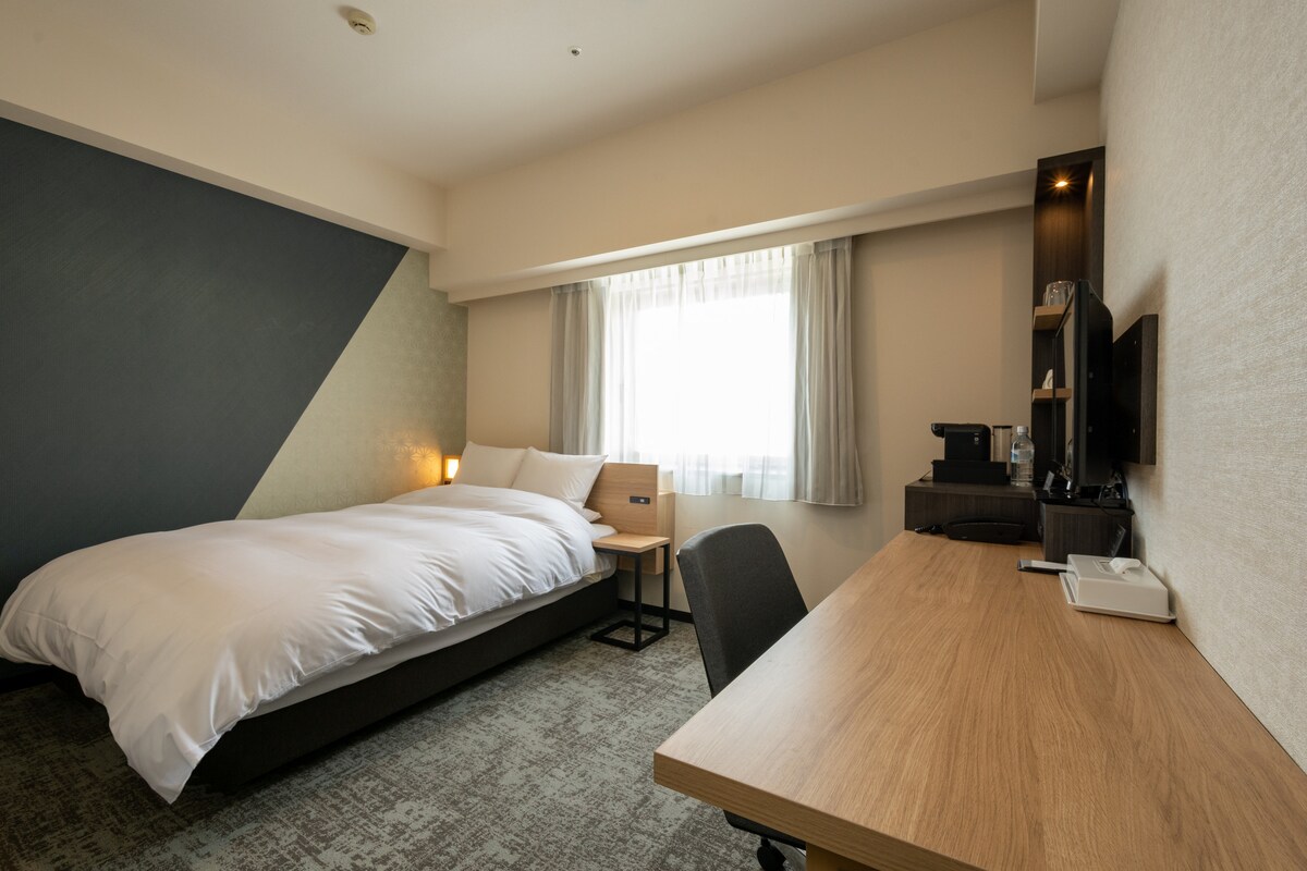 A spacious room features a large bed with a clean white comforter, complemented by a side table and a desk. A window allows natural light to fill the space, and a subtle color palette is present on the walls, enhancing the calm environment.