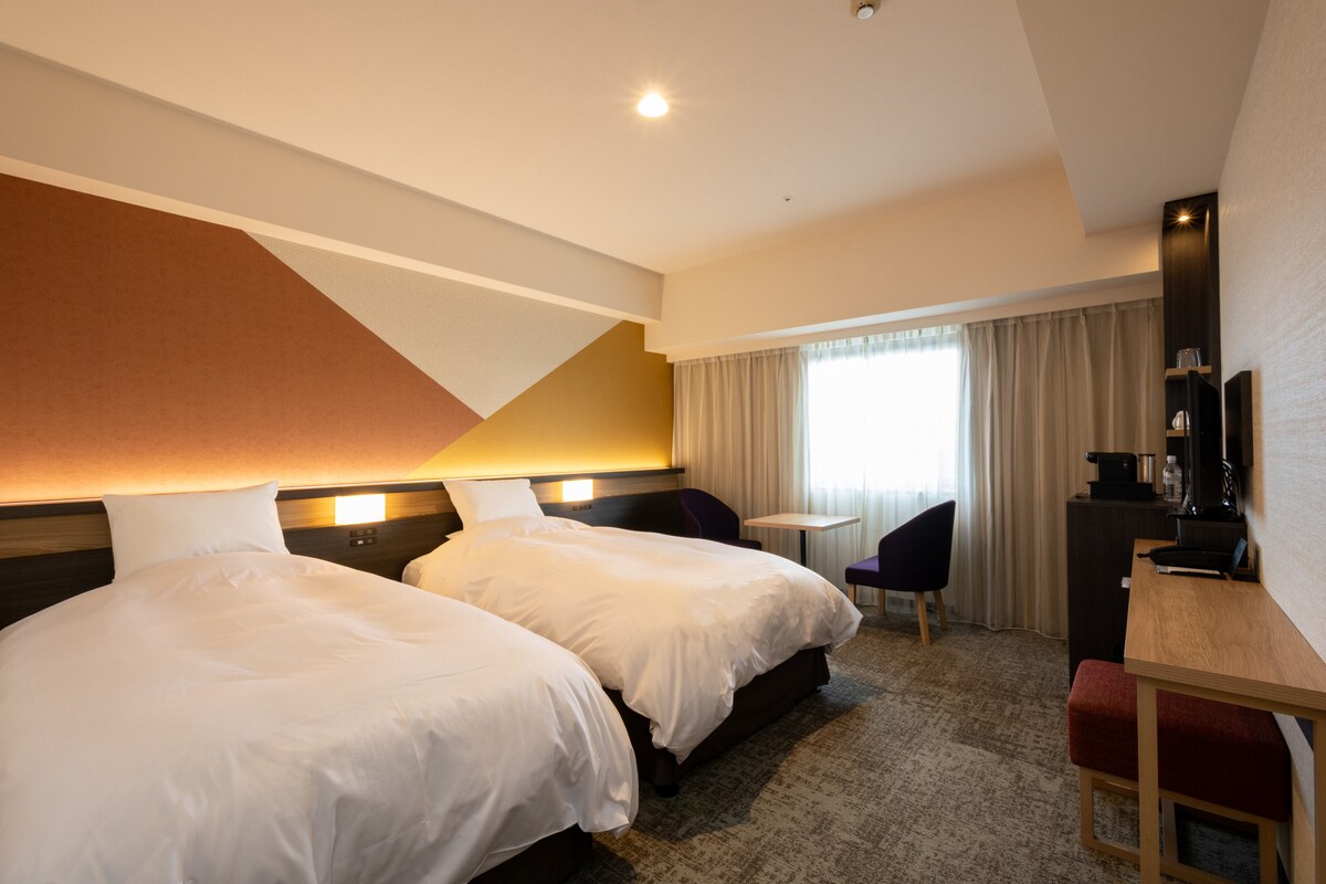 A well-appointed room features two spacious beds, each with white bedding, positioned beside a window dressed in light curtains. A small table and two chairs are visible, along with a coffee station and a flat-screen TV against a modern wall design.