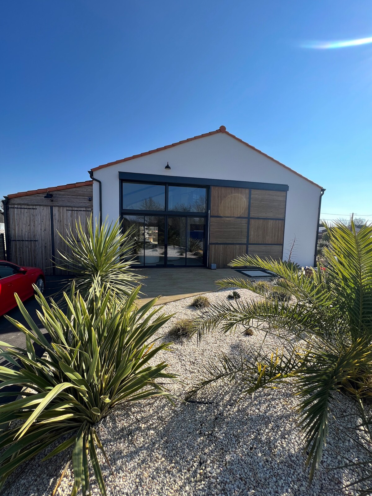 A contemporary villa features a striking façade with large glass windows and wooden accents, inviting natural light. Surrounding foliage consists of palm plants and a gravel garden, enhancing the outdoor aesthetic. The clear blue sky creates a bright and open atmosphere.