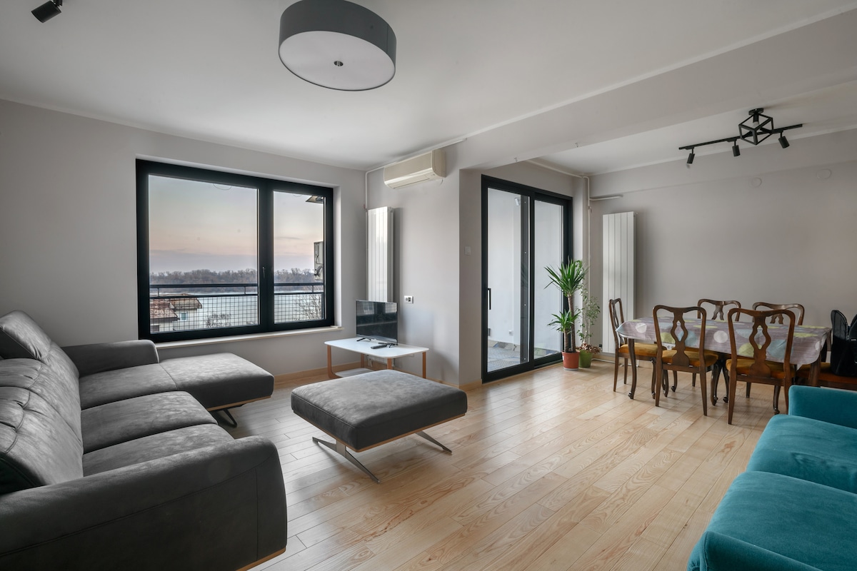 A spacious living area features large windows offering views of the waterfront. A gray sectional sofa and an ottoman provide comfortable seating. A dining table for six is positioned adjacent, while a potted plant adds a touch of greenery to the space.