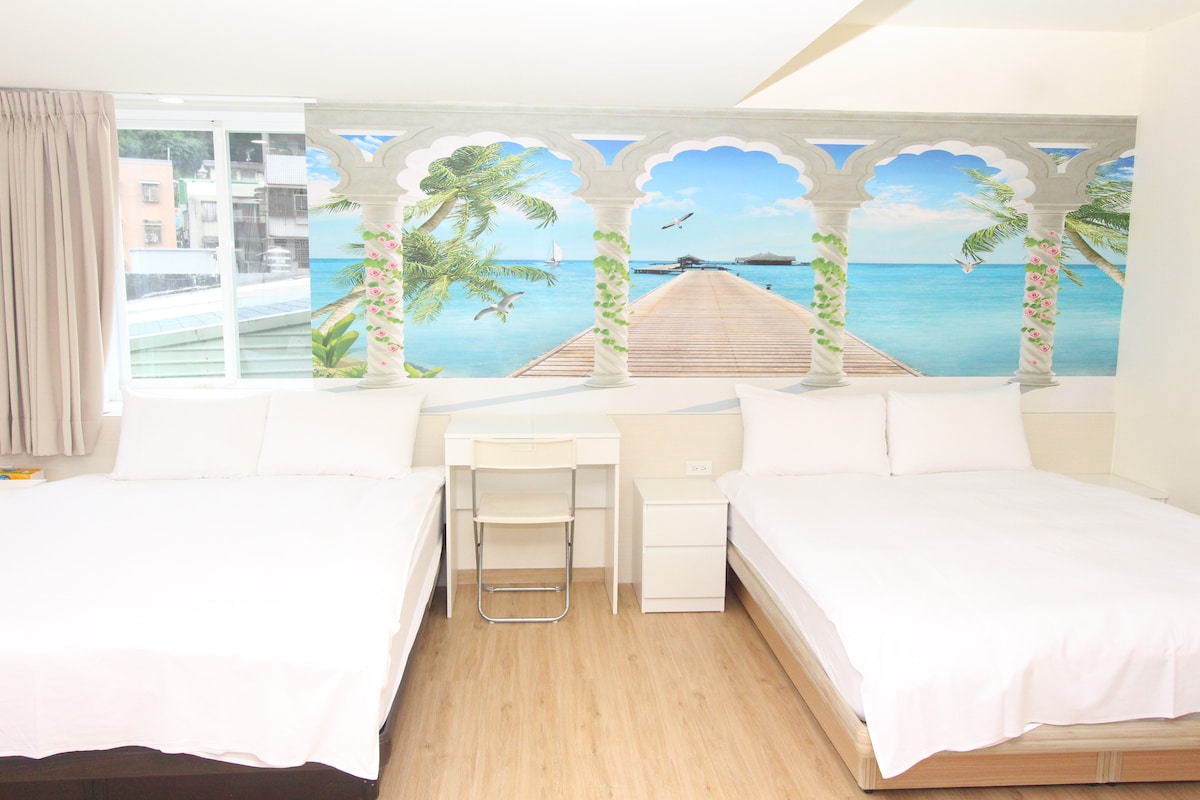 The room features two spacious beds with white linens, positioned against a vibrant mural depicting a serene pier over calm waters. Natural light enters through large windows, and a small desk with a chair is placed between the beds, offering a functional workspace.