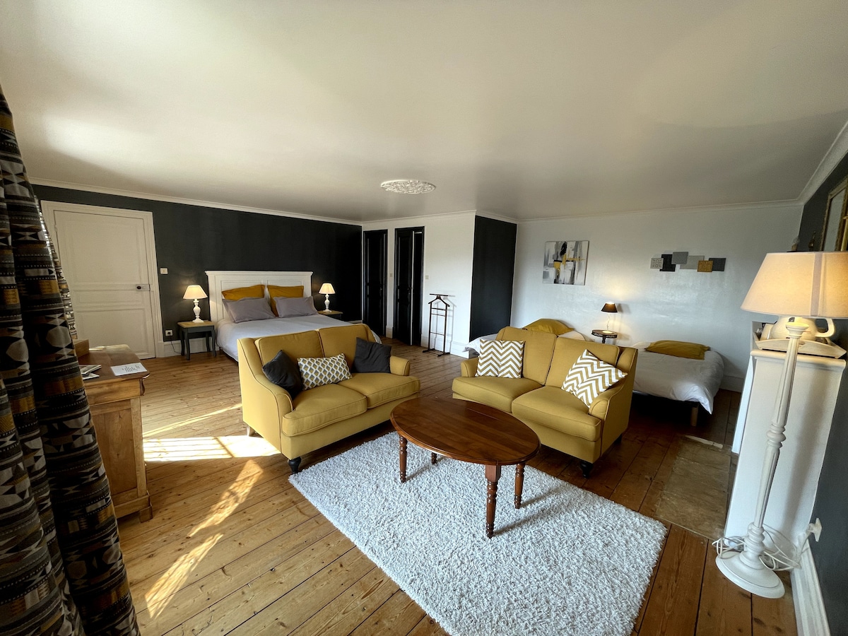 A spacious room features a comfortable seating area with a yellow sofa and a round wooden table. A large bed is positioned against a dark wall, while a second bed is situated nearby. Natural light floods in through a nearby window, illuminating the polished wood floor.
