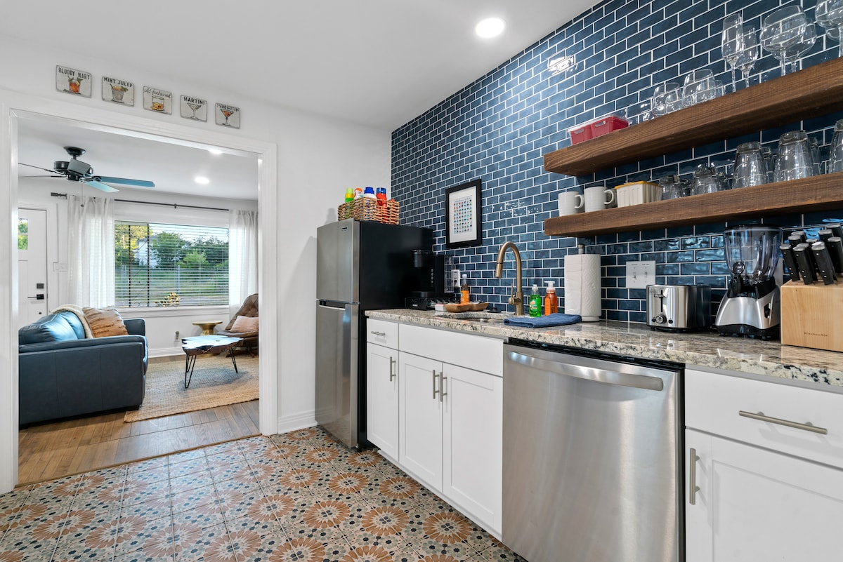 A modern kitchen is highlighted with blue tiled walls and a granite countertop. Stainless steel appliances, including a refrigerator and dishwasher, are visible. Open shelving displays glassware and kitchen essentials. A doorway leads to a living area with comfortable seating and natural light from large windows.