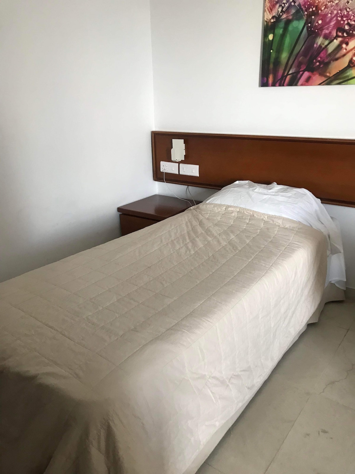 A single bed covered with a light-colored quilt is positioned against a wooden headboard. A nightstand with power outlets is located beside the bed. Neutral walls and floor create a calm backdrop.