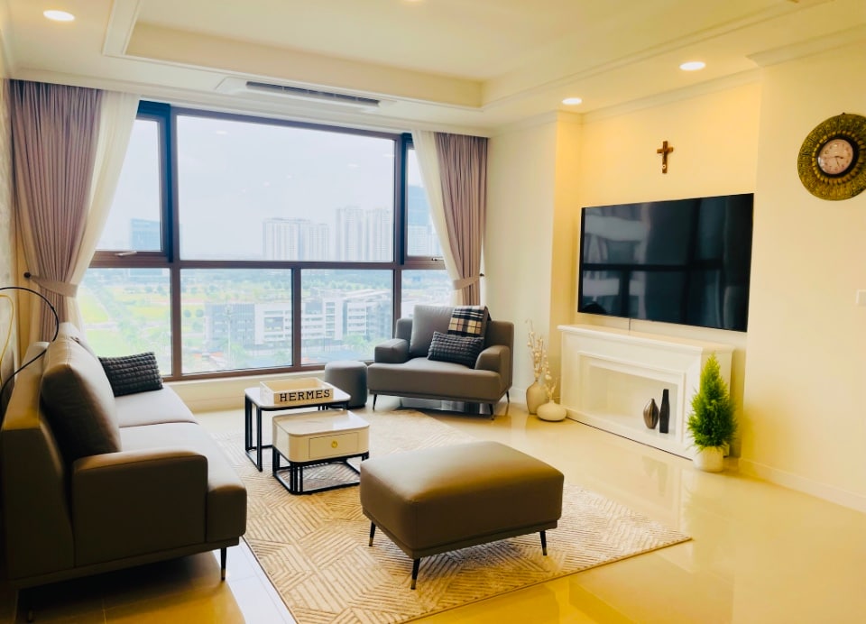 A bright living area is illuminated by natural light from a large window, offering views of the surrounding landscape. Comfortable gray sofas and a matching ottoman are arranged around a coffee table. A flat-screen television is mounted on the wall, complemented by decorative elements.