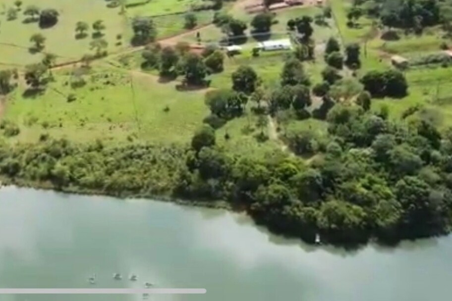 Aerial view of a serene landscape featuring a body of water, surrounded by lush greenery and trees. Several structures are visible in the distance, along with a shaded pathway leading through the natural surroundings.