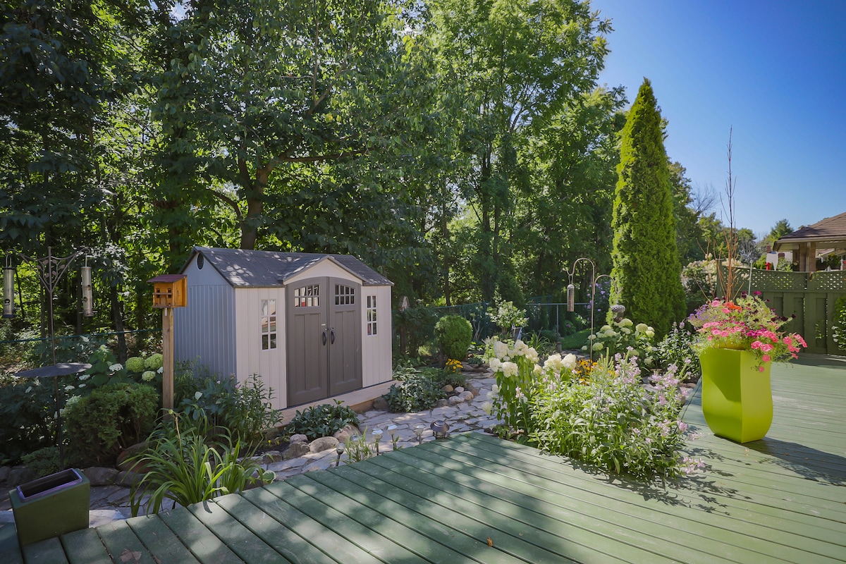 A landscaped backyard features a small garden shed with double doors, surrounded by lush greenery and colorful flowers. A stone pathway winds through the garden, complemented by a green deck and vibrant planters that add a touch of color.