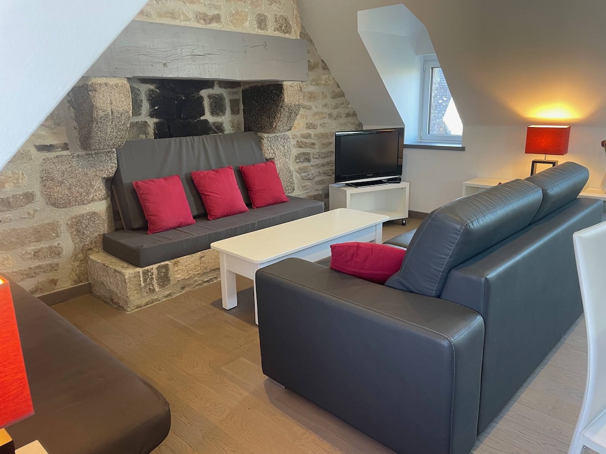 A spacious living area features a stone fireplace and two comfortable couches, one adorned with red accent pillows. A television is positioned on a stand, complemented by a white coffee table. Natural light is welcomed through slanted windows, enhancing the modern decor.