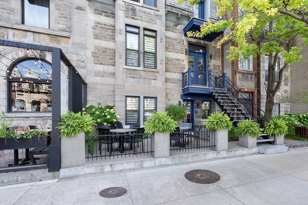 A charming exterior view showcases a layered stone building complemented by blue accent details. A staircase leads to a second floor, surrounded by lush greenery in planters and outdoor seating arranged on a patio, creating an inviting atmosphere alongside the street.