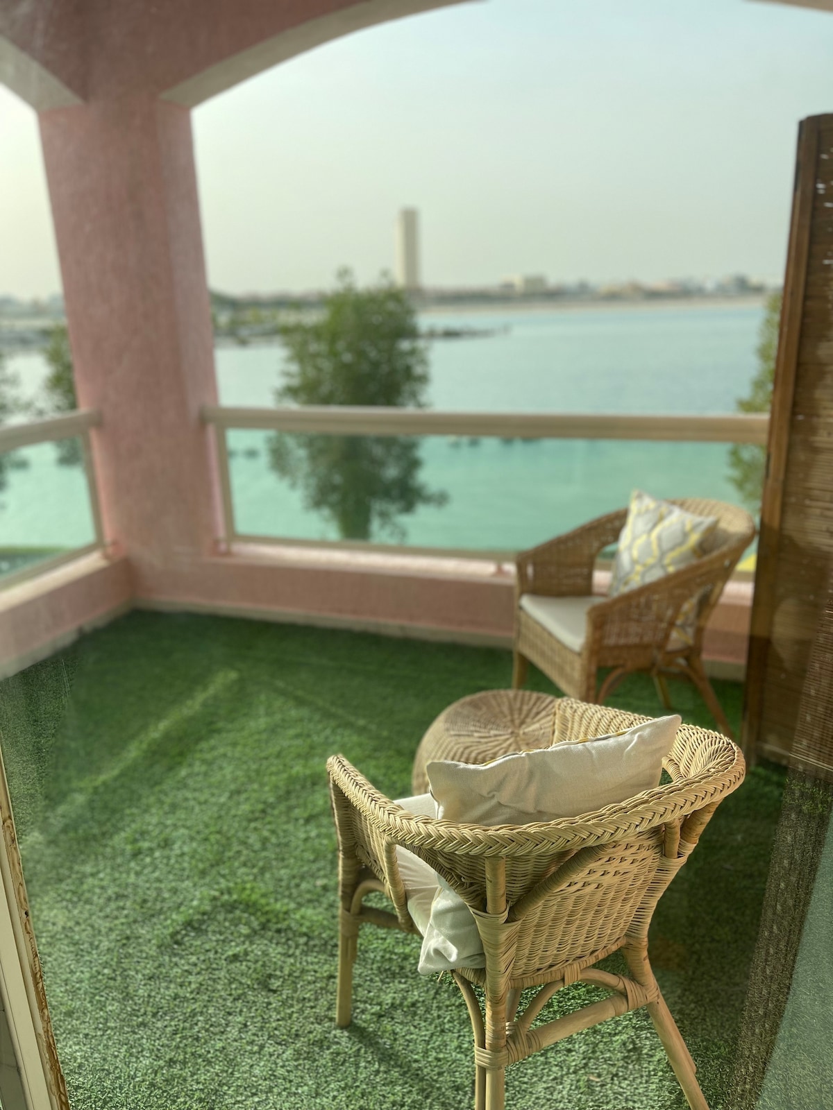 An outdoor seating area is featured with two wicker chairs and cushions positioned on a green carpeted surface. The view overlooks the water, with a distant building visible on the shoreline.