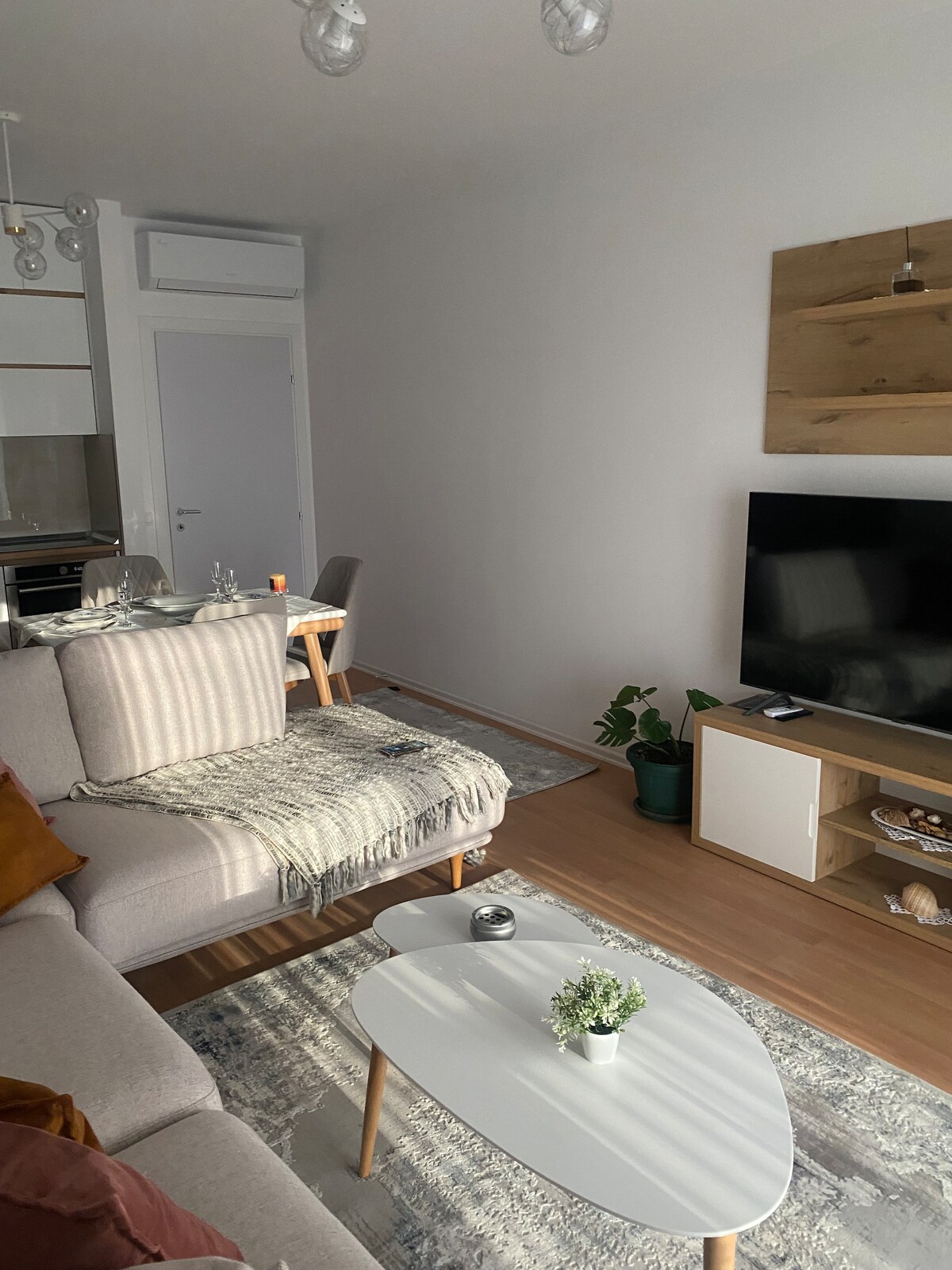 A modern living space is depicted, featuring a light grey sofa adorned with throw pillows and a throw blanket. A small white coffee table sits in front, accompanied by a decorative plant. A television is positioned on a cabinet, with a dining area visible in the background.