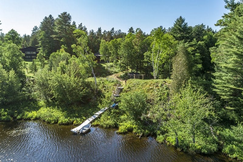 The image captures a serene waterfront setting along Stony Creek Ponds, showcasing a private dock leading into the water. Lush greenery surrounds the area, with a mix of trees and shrubs providing natural privacy. The distant outline of a building is visible among the foliage.
