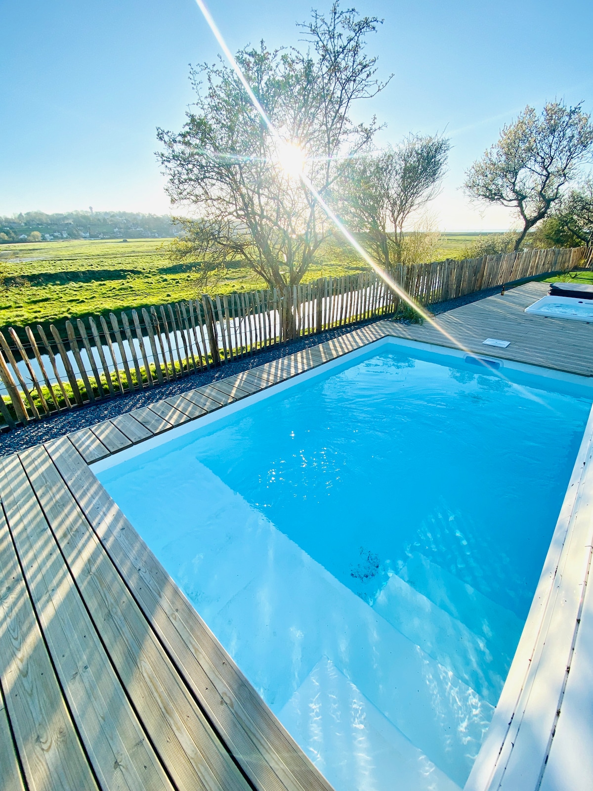 A private pool is set within a wooden deck, surrounded by greenery and scenic views. Sunlight reflects on the water's surface, creating a serene ambiance. The area is enclosed by a wooden fence, providing a sense of privacy and tranquility.