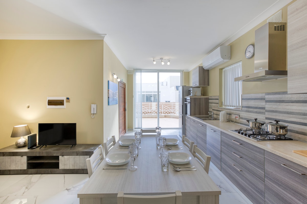 The modern kitchen space features a long wooden dining table set for six, accompanied by stylish chairs. An open layout connects to a living area with a television and large windows that allow natural light. Neutral tones enhance the airy atmosphere.