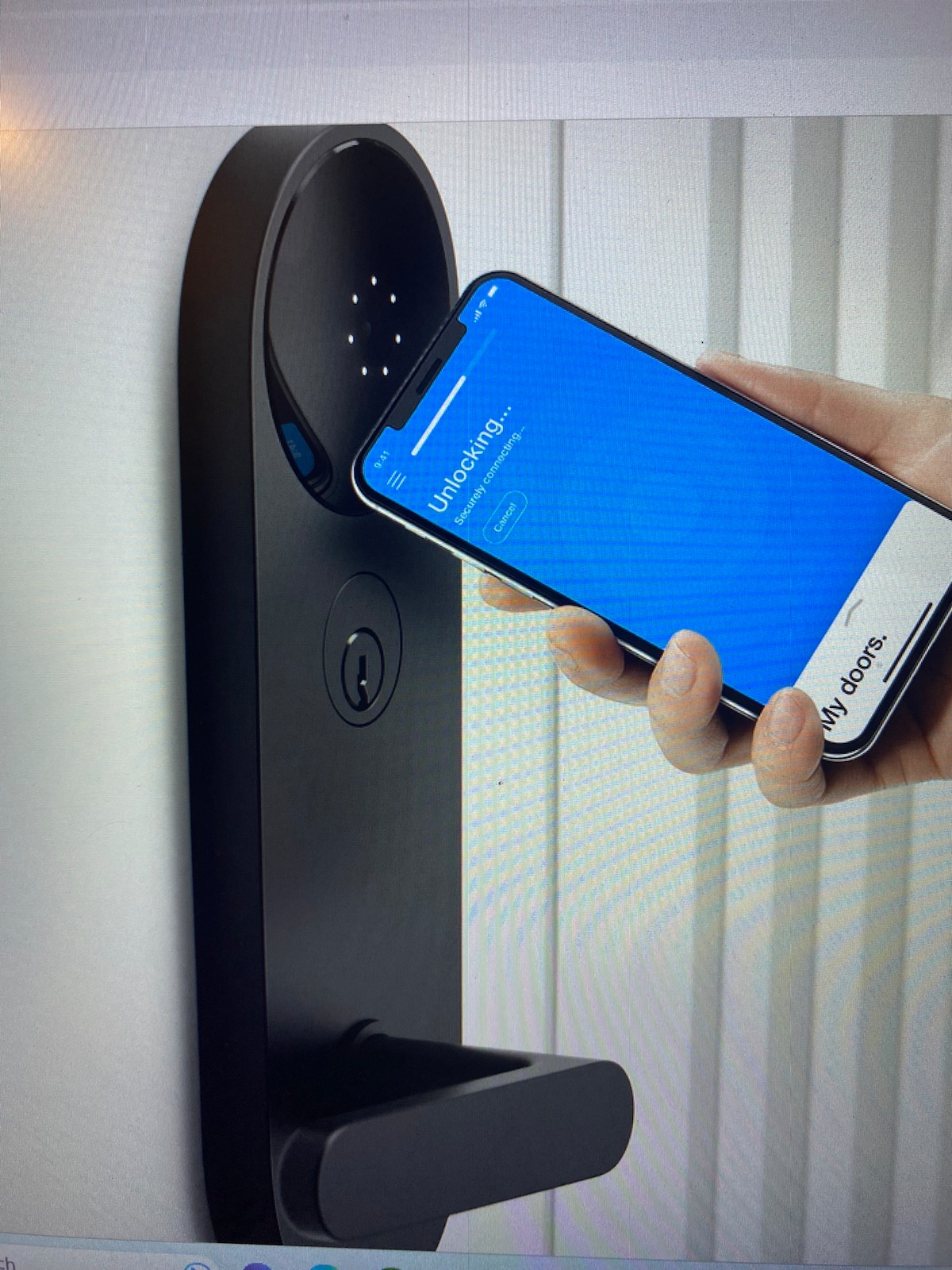 A smart lock is shown being activated by a smartphone. The lock is sleek and modern, featuring a black finish with illuminated numbers indicating its technological features. The background consists of vertical blinds, adding subtle texture to the image.