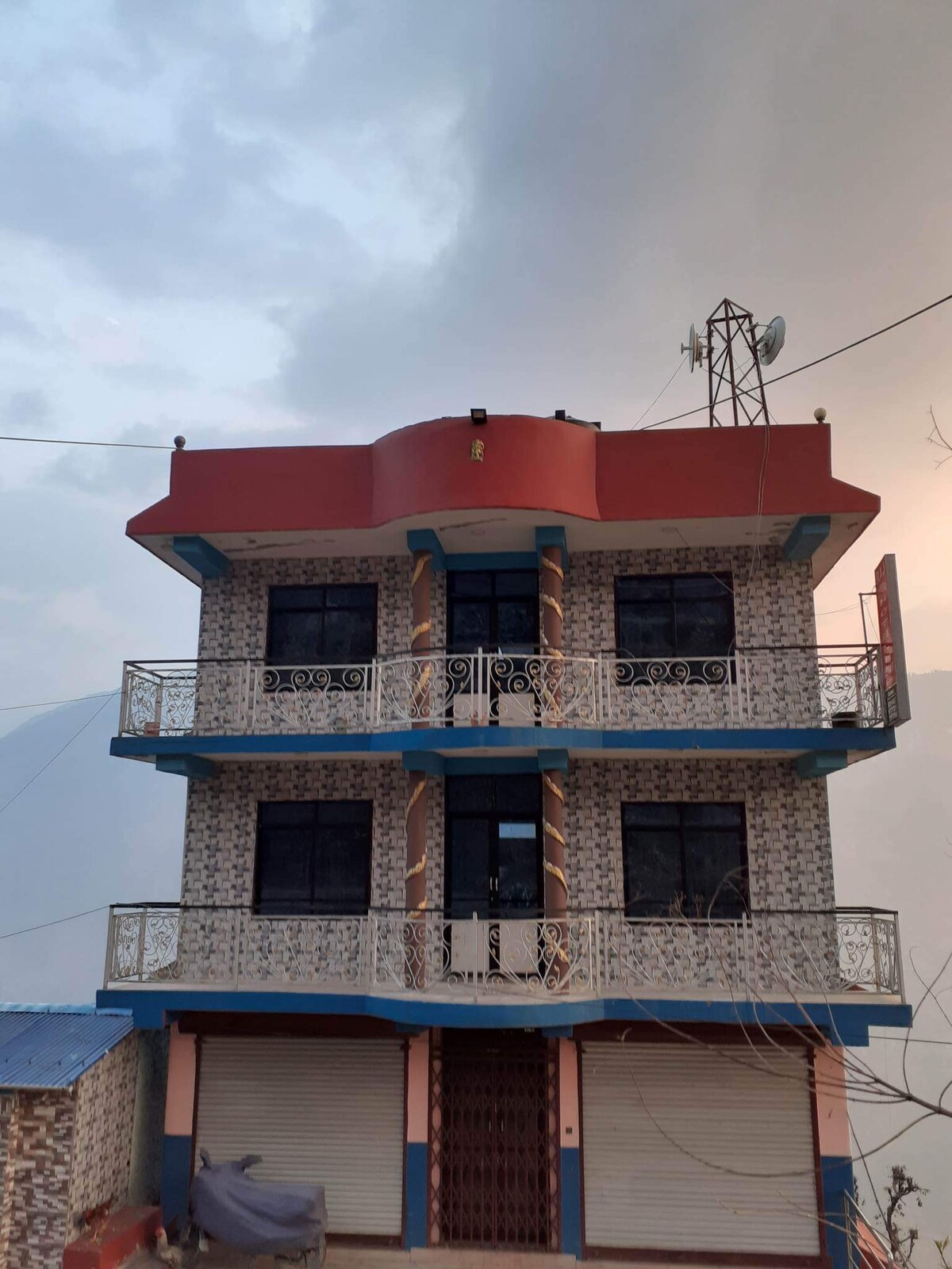 A three-story building is presented with a distinctive red rooftop and blue accents. The facade is adorned with patterned tiles and large windows, allowing ample light to enter. A balcony borders the second and third floors, providing views of the surrounding area.