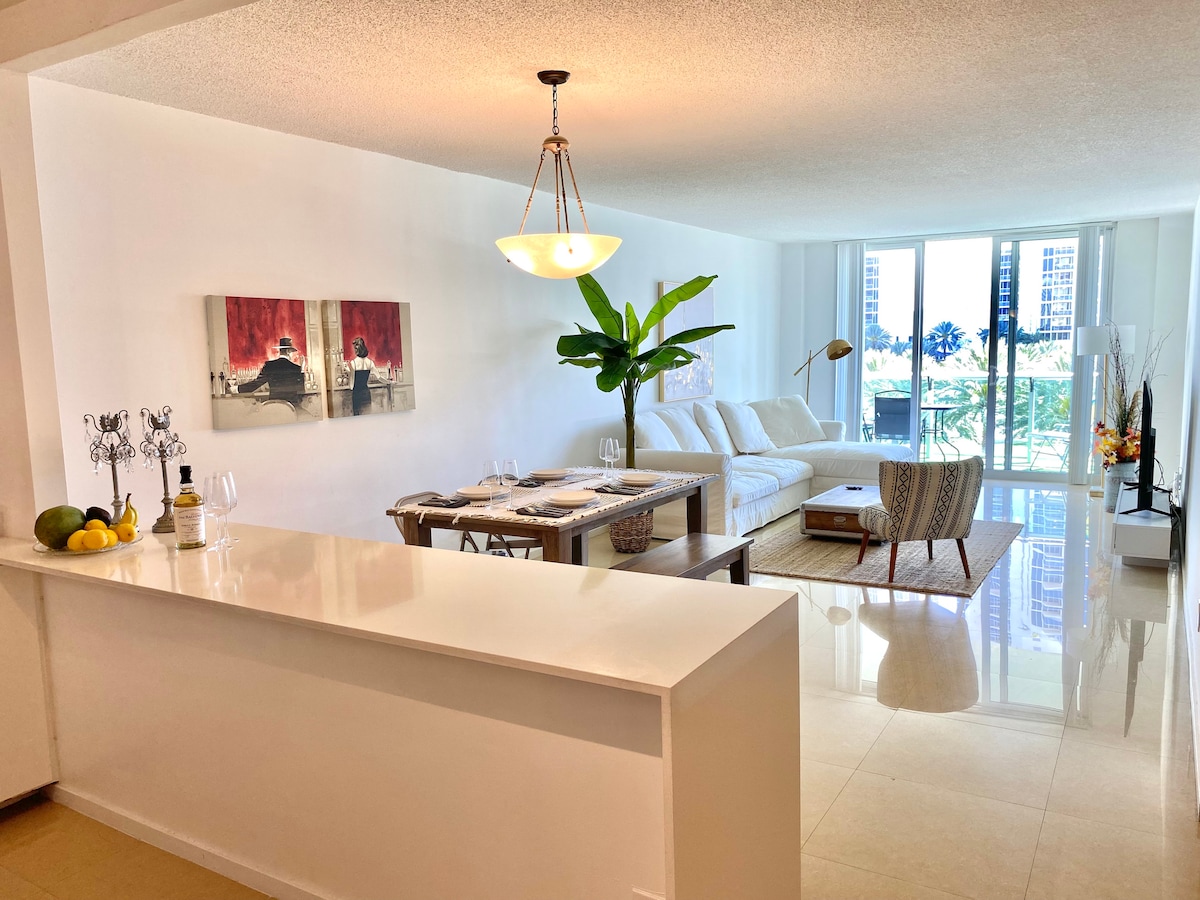 A modern living space is depicted, featuring a light-colored sofa paired with a patterned chair. A glass dining table set for two is visible, accompanied by a fruit display. Natural light fills the room through large sliding doors, showcasing views of the surrounding greenery.