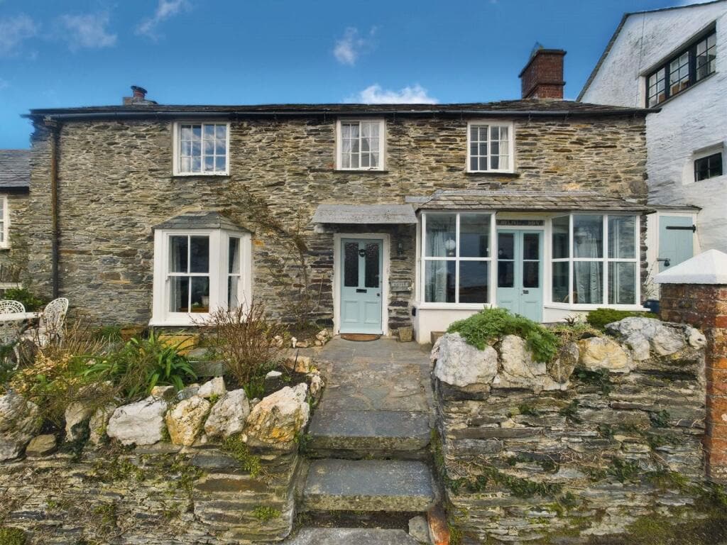 A charming stone cottage is featured, showcasing a classic design with light blue doors and large windows. The entrance is framed by natural stone landscaping and greenery, providing a welcoming facade. A pathway leads up to the front door, inviting guests to explore further.