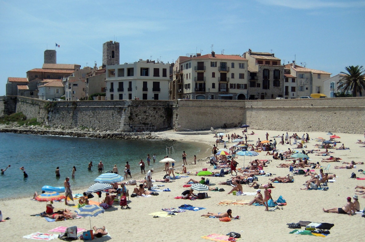 A sandy beach is filled with sunbathers and umbrellas, while crystal-clear waters gently lap at the shore. Historic buildings with a Mediterranean style rise in the background, offering a glimpse of the old town. A palm tree adds a tropical touch to the scene.