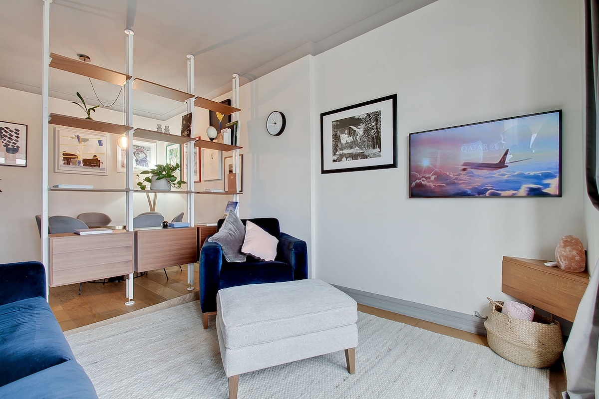 An open plan living area showcases a cozy blue armchair and a light-colored ottoman on a large woven rug. Modern shelving acts as a divider, while framed artwork adorns the walls. A television mounted on the wall adds to the contemporary furnishings.