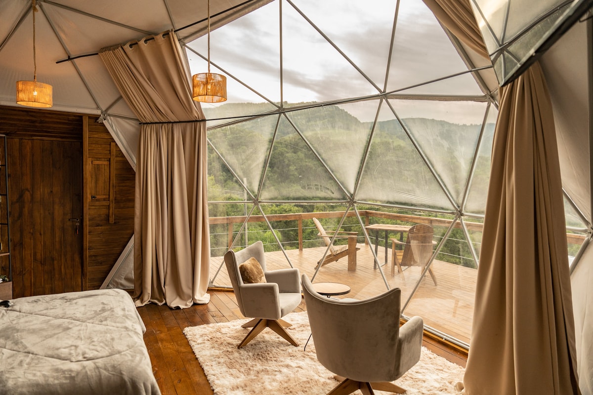 A geodesic dome interior offers a spacious ambiance with large windows framing lush green views. A comfortable seating area is present with two chairs and a small table. Natural light filters in through the translucent panels, complemented by warm wooden accents.