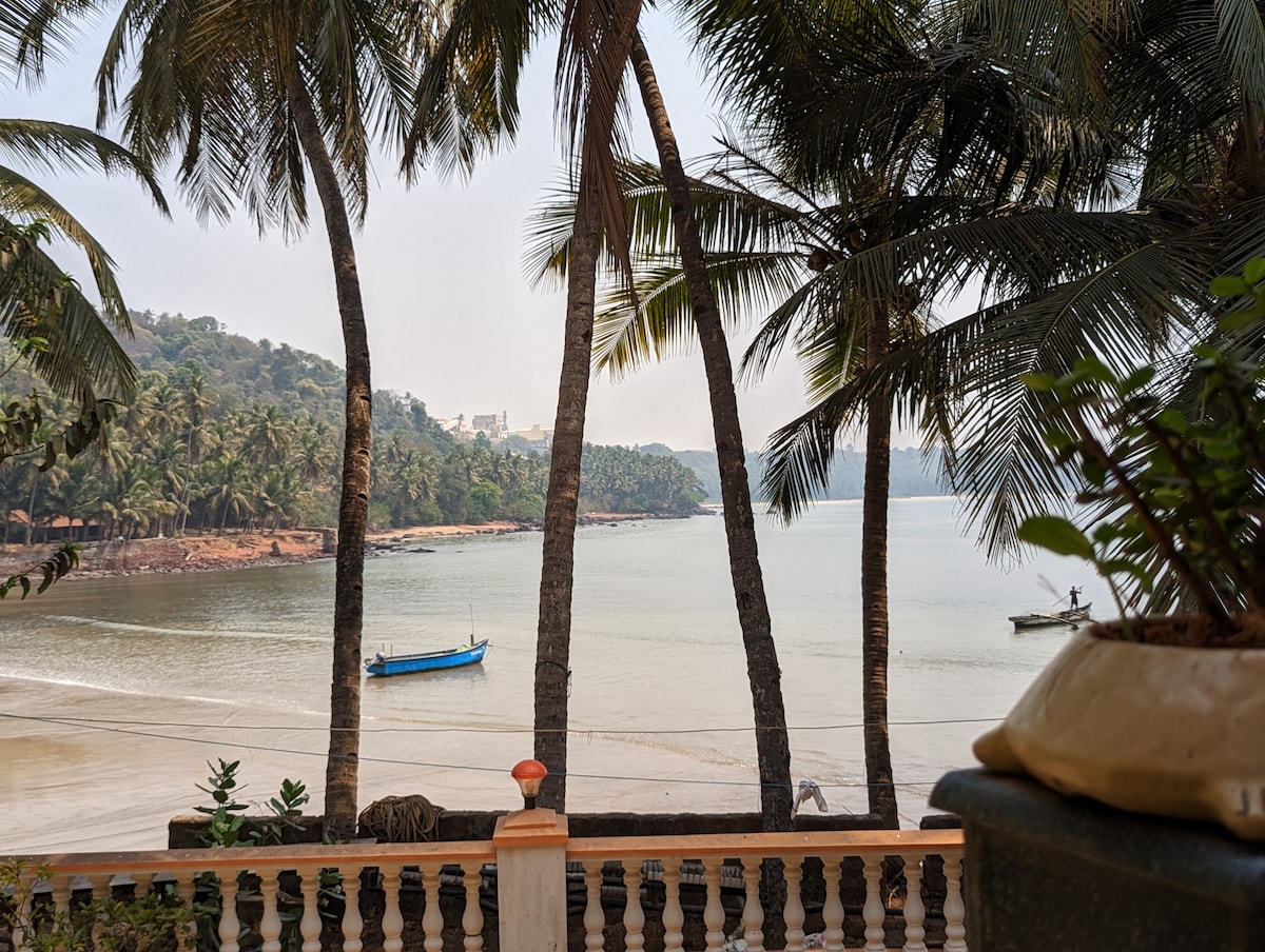 A serene beach view features gentle waves lapping against the shore, framed by swaying palm trees. In the distance, fishing boats can be seen on the water, enhancing the tranquil atmosphere of the landscape.