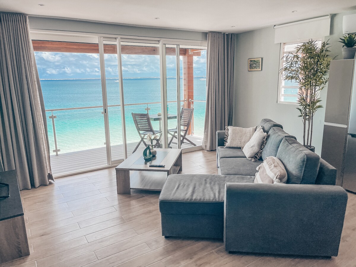 A living area features a comfortable sectional sofa paired with a coffee table. Floor-to-ceiling glass doors open to a private balcony, revealing a sweeping view of the sea. A small dining set with two chairs is positioned near the balcony, enhancing the open feel.