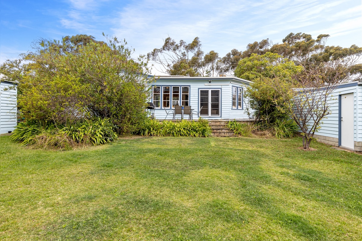 The exterior view showcases a charming cottage surrounded by lush greenery and well-maintained grass. Steps lead up to the entrance, which features multiple windows and a small outdoor seating area. Nearby, storage sheds are visible, framed by hedges and shrubbery.