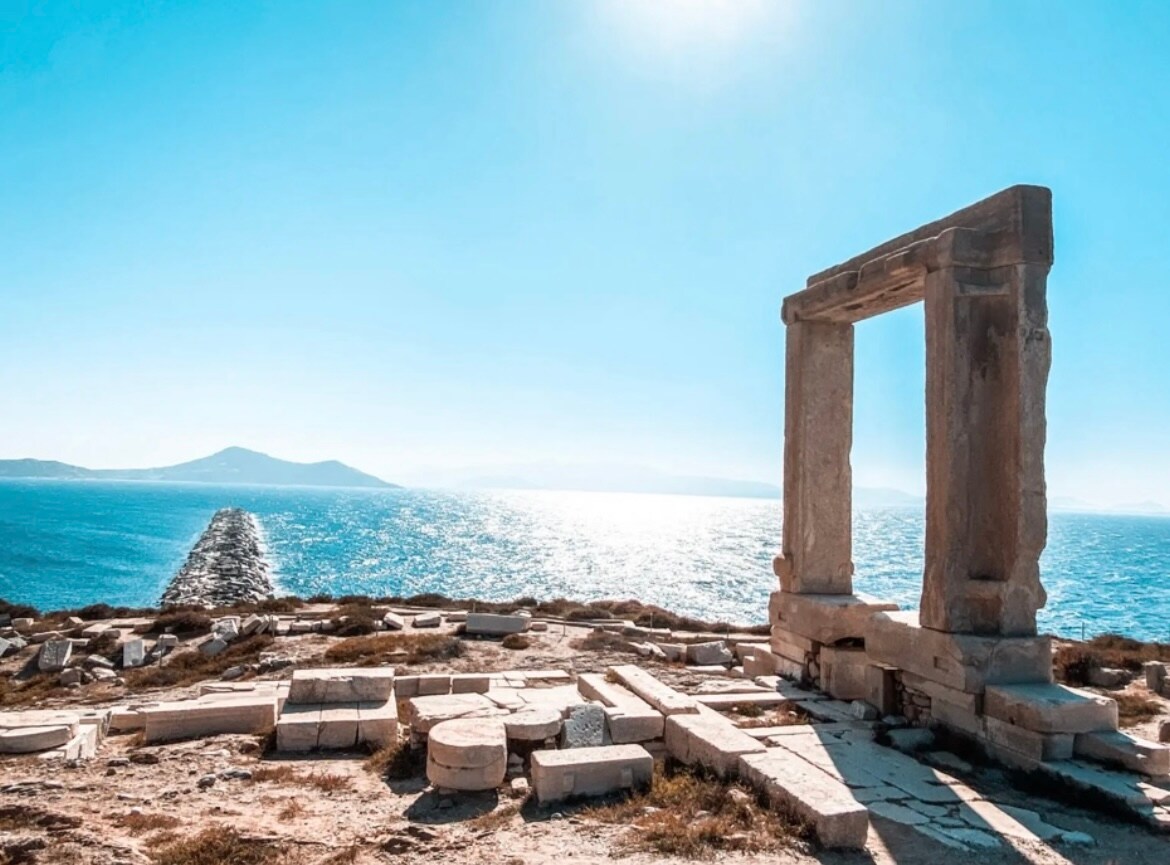 A historic stone entrance stands prominently against a bright blue sky and the shimmering sea. Ruins of broken columns and fragments are scattered across the landscape, leading towards a distant rocky jetty extending into the water.