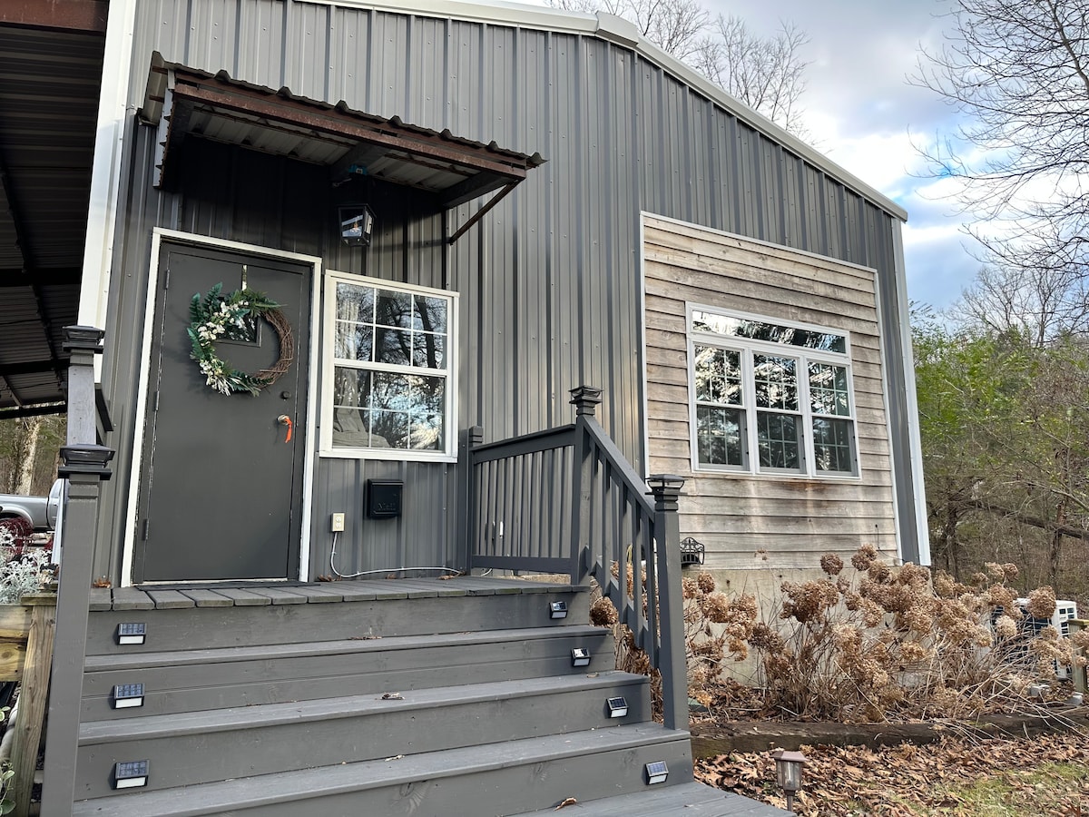 The entrance of the cottage is framed by wooden steps leading to a dark door adorned with a festive wreath. Large windows are visible on either side, reflecting the natural surroundings, while a covered porch area provides shelter from the elements.