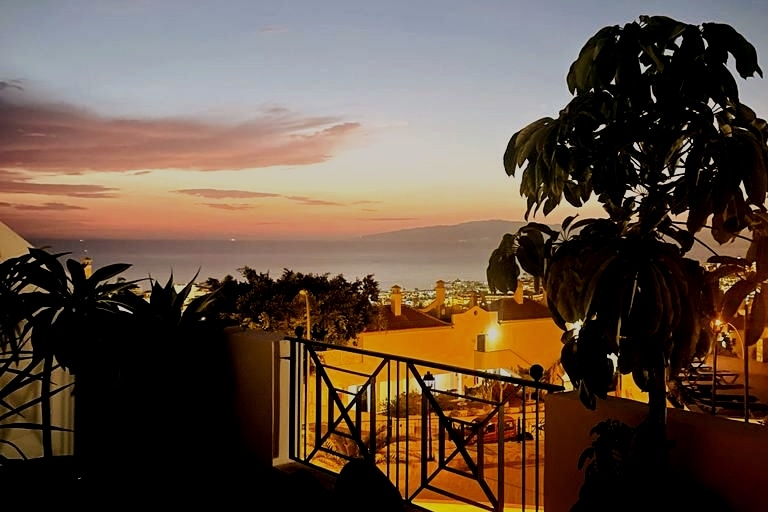 A balcony view captures the breathtaking sunset over the ocean, with silhouettes of palm trees framing the scene. The coastline glows under warm evening light, highlighting the tranquil environment of Costa Adeje.