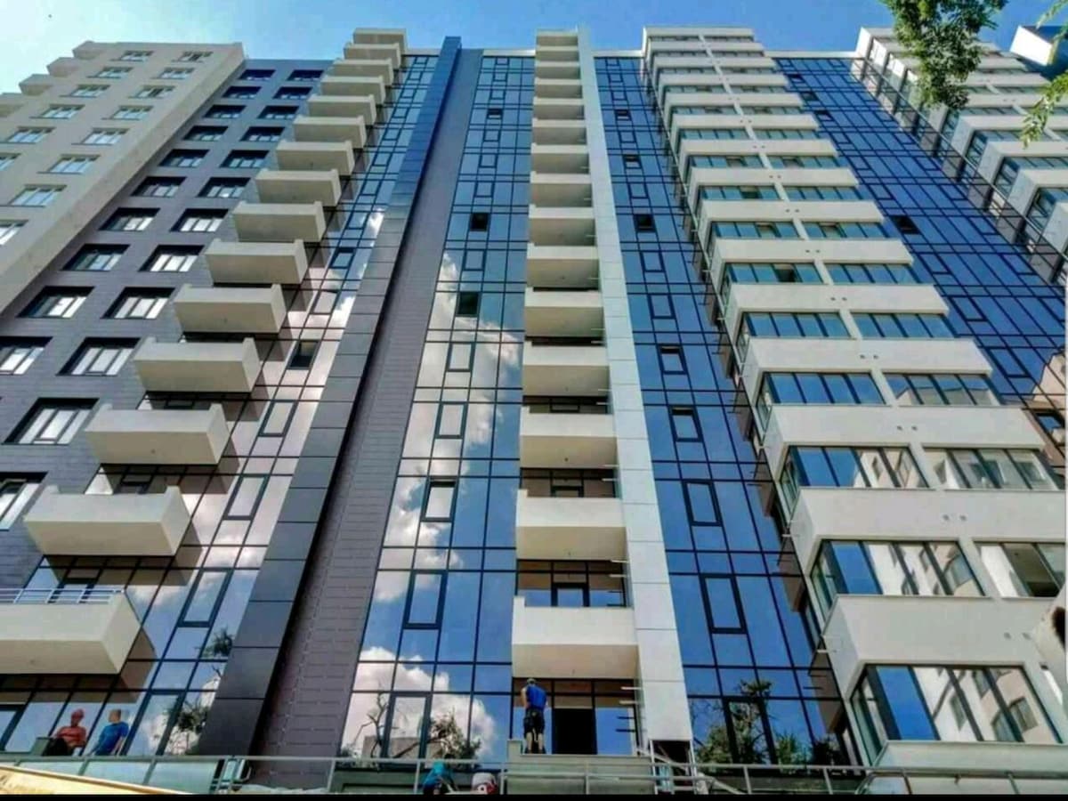 The image depicts the exterior of a modern apartment building featuring multiple floors with large glass windows and a mix of light and dark facade colors. Balconies are visible on several levels, enhancing the contemporary architectural design.