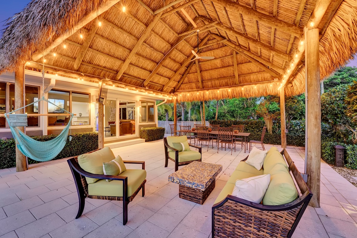 A spacious outdoor patio features comfortable seating arrangements, including a woven lounge set with cushions. A large, thatched roof provides shelter, with string lights adding a soft glow. A hammock is gently swaying nearby, enhancing the relaxing ambiance of the garden setting.