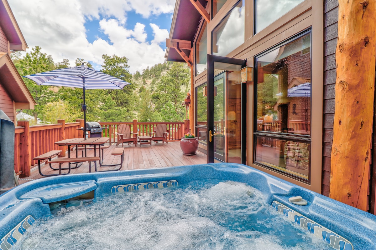 A bubbling hot tub is positioned on the spacious deck, surrounded by wooden railings. An umbrella shades a nearby seating area with a picnic table. Large glass doors lead into the cabin, allowing natural light to illuminate the interior.