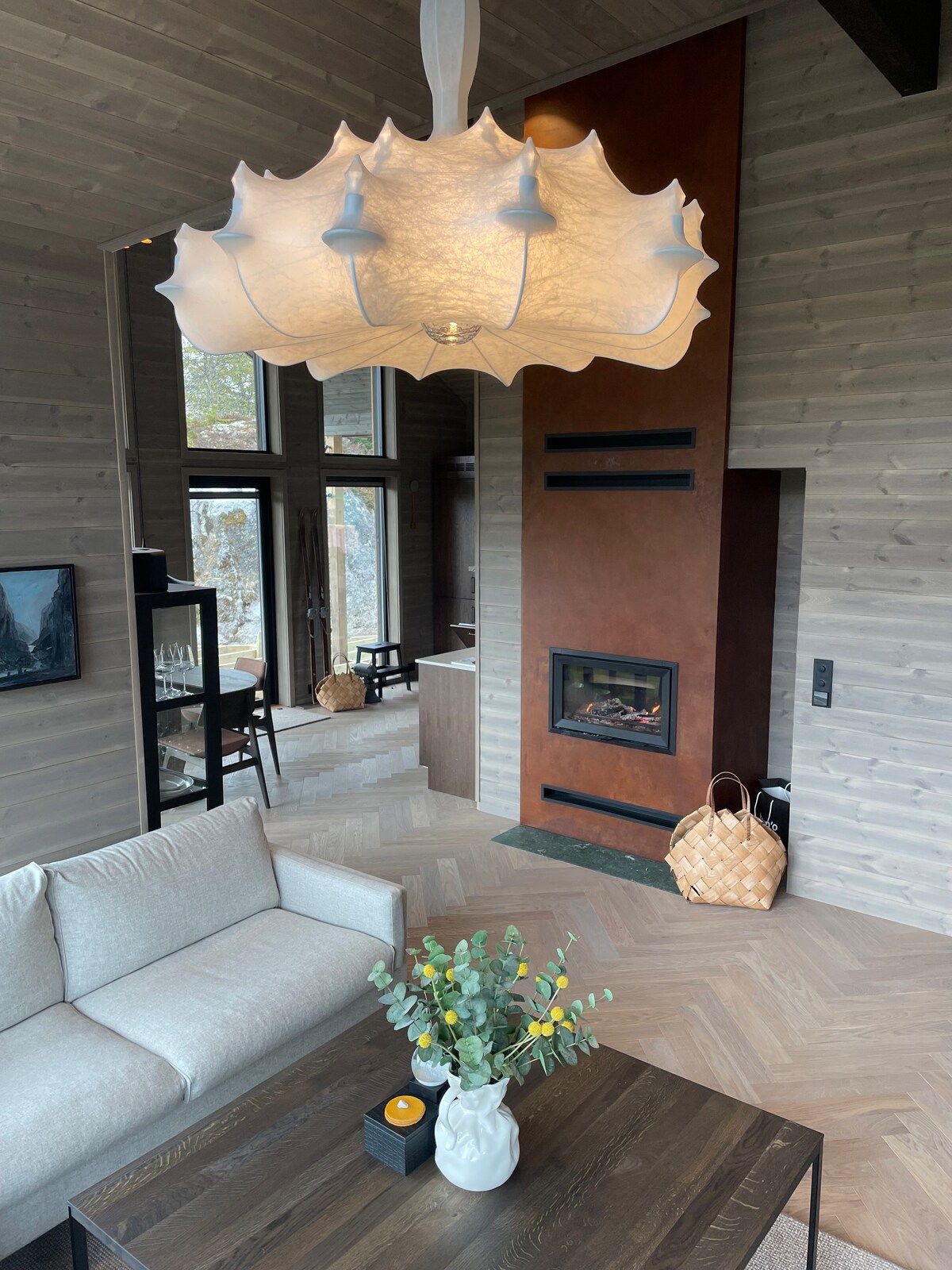 A spacious living area features a soft, textured chandelier hanging elegantly above a minimalistic coffee table. Large windows provide natural light, while a modern fireplace adds a cozy touch to the room's design. Warm wooden tones enhance the inviting atmosphere.