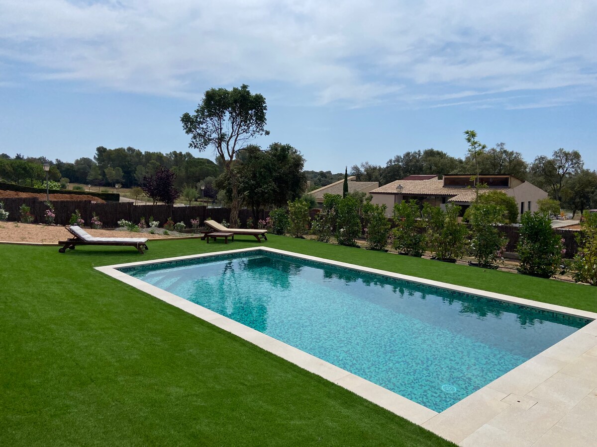 A spacious outdoor area highlighted by a shimmering pool surrounded by well-maintained grass. Two lounge chairs are positioned nearby, offering a spot for relaxation. Lush greenery and flowering plants frame the pool, contributing to the serene ambiance.