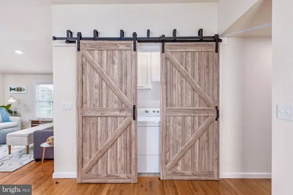 A set of double barn doors, featuring a rustic wood finish, provides access to the laundry area. The doors are mounted on a black metal track against a light-colored wall. The hardwood floor extends into the room, enhancing the clean and modern look.