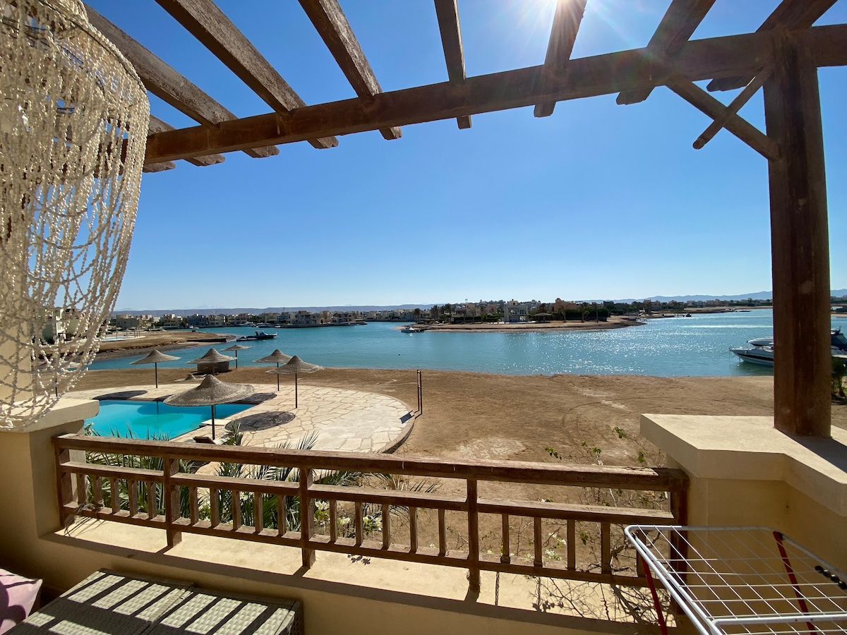 A scenic view features serene waters framed by a wooden pergola. The image captures the beach area and pool below, with sunlit surroundings and a clear blue sky. The space offers a sense of tranquility and connection to the natural landscape.
