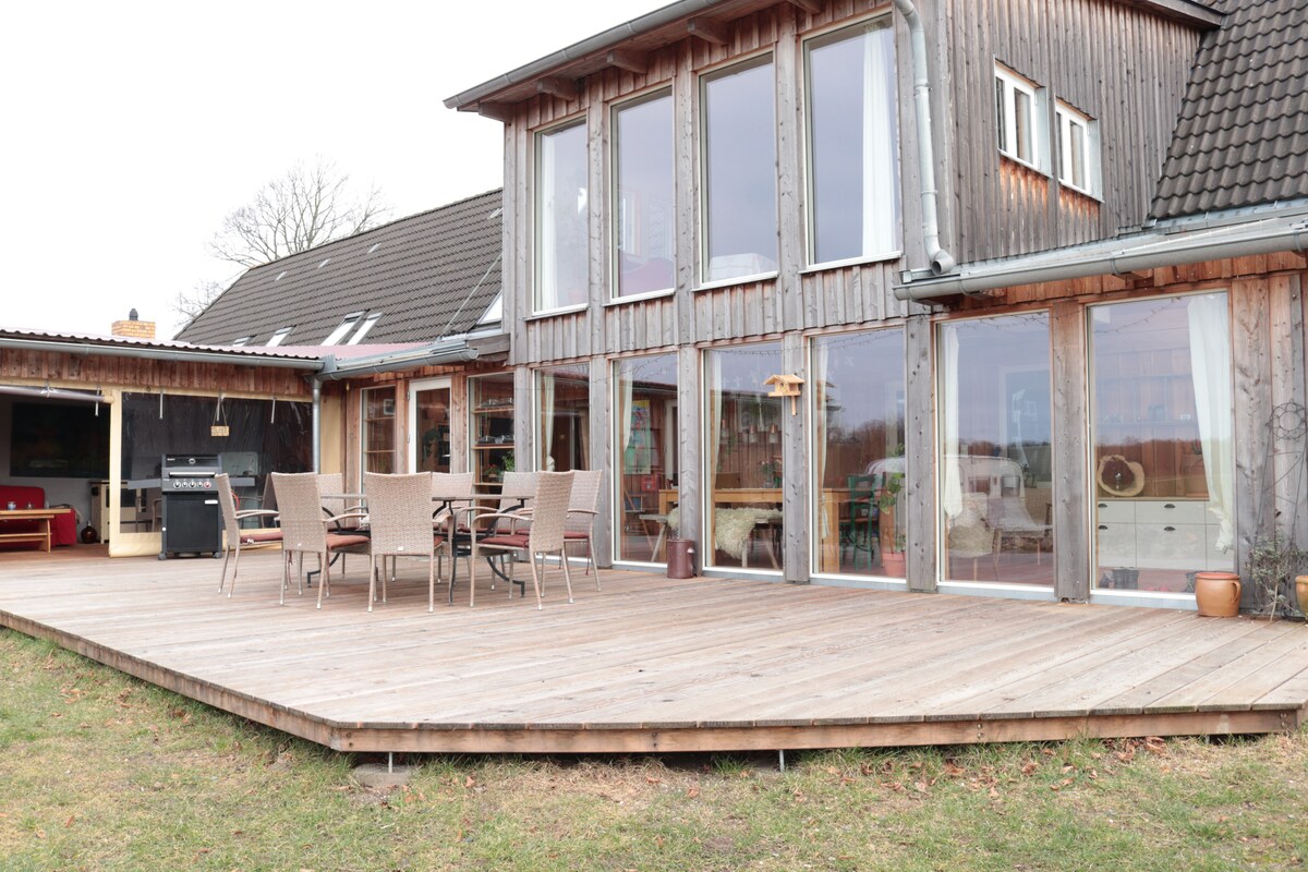 A wooden terrace extends from the house, featuring a dining table surrounded by six chairs. Large glass windows create a seamless connection with the outdoor space. The entrance to an adjoining covered area is visible, enhancing the open atmosphere of the environment.
